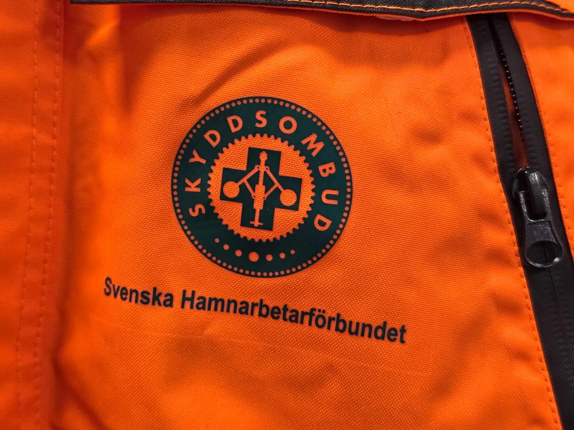 Skyddsombudsmärke med texten Svenska Hamnarbetarförbundet på en orange arbetsjacka