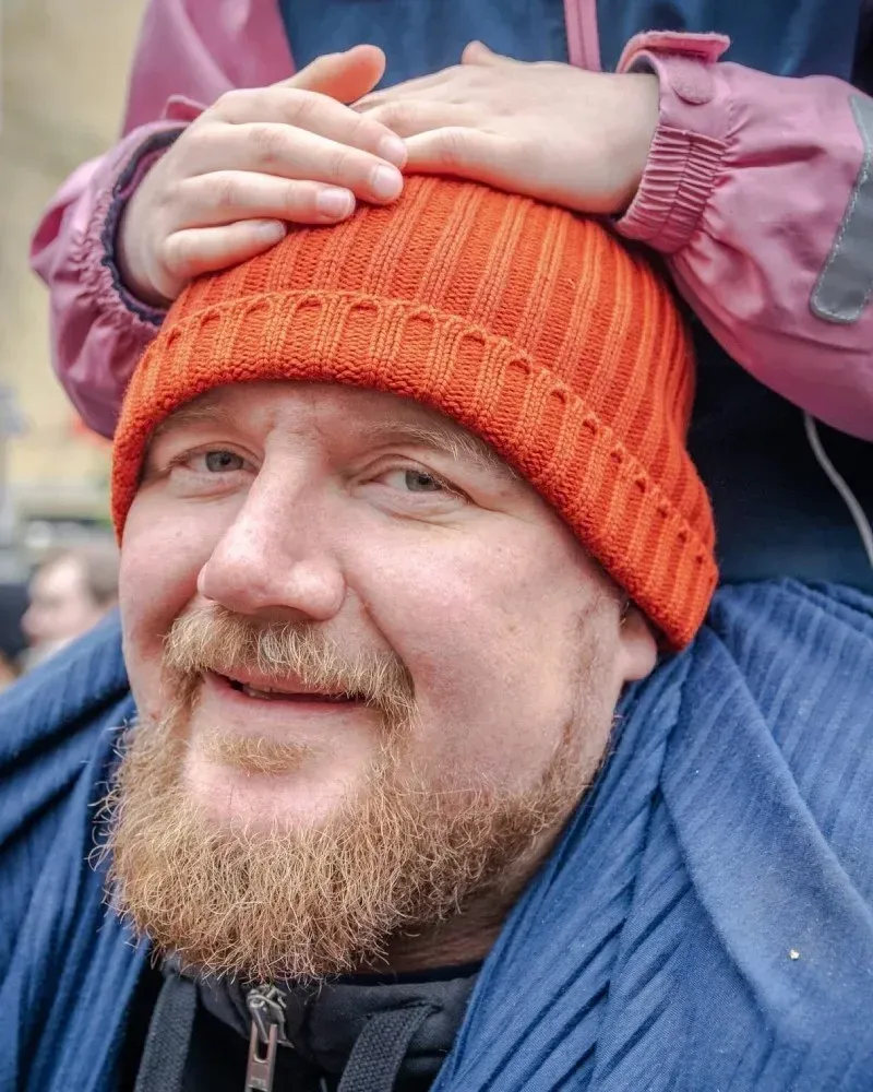 Man med skägg och orange mössa som bär ett barn på axlarna, barnets händer vilar på hans huvud