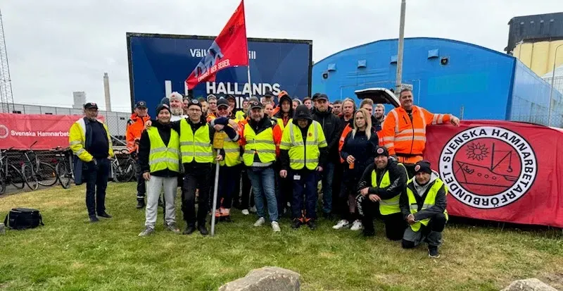 Hamnarbetare i gula och orange varselvästar samlade utomhus med Hamnarbetarförbundets röda fana och banderoll