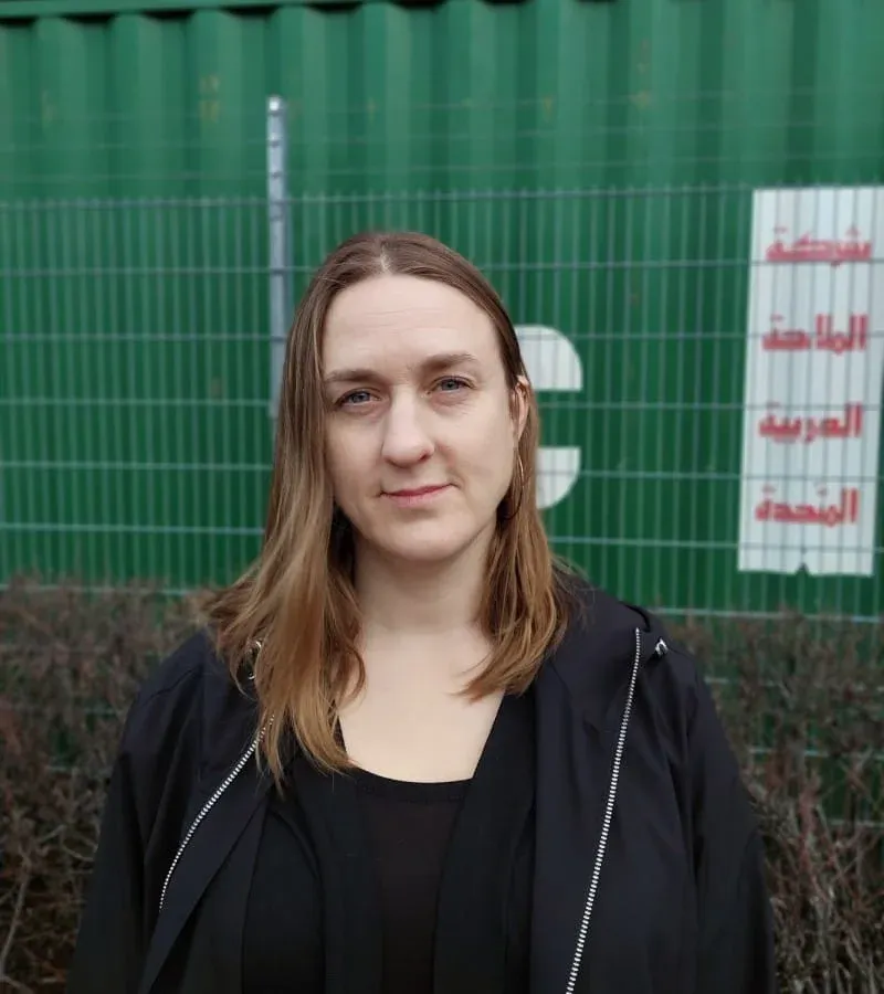 Porträtt av en kvinna i svart jacka framför ett grönt staket och en container